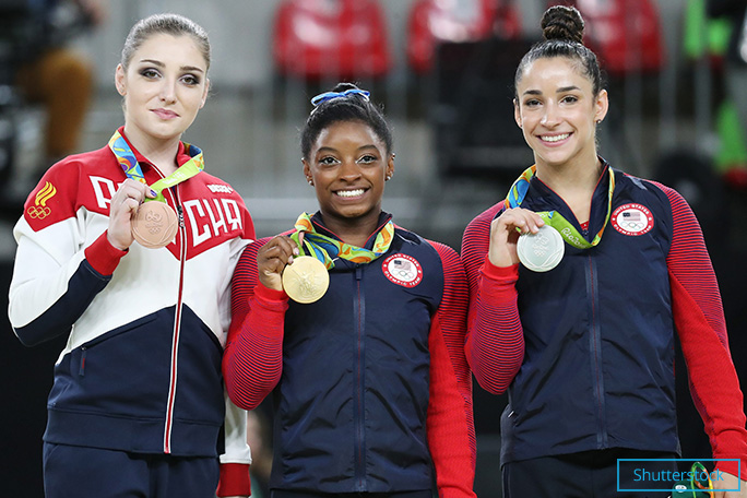 2016年リオデジャネイロオリンピックで個人総合の金メダルを見せるバイルズ。隣は銀メダリストのアレクサンドラ・レイズマンと銅メダリストのアリーヤ・ムスタフィナ。